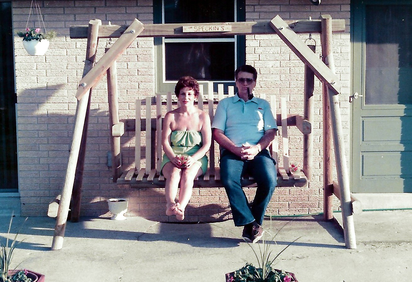 Couple sits on a large wooden outdoor swing in front of a brick house, with potted plants nearby.