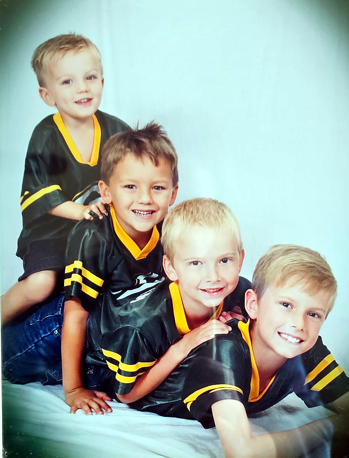 Four young boys in matching black and yellow sports jerseys posing together, stacked on a white surface with a light blue backdrop.