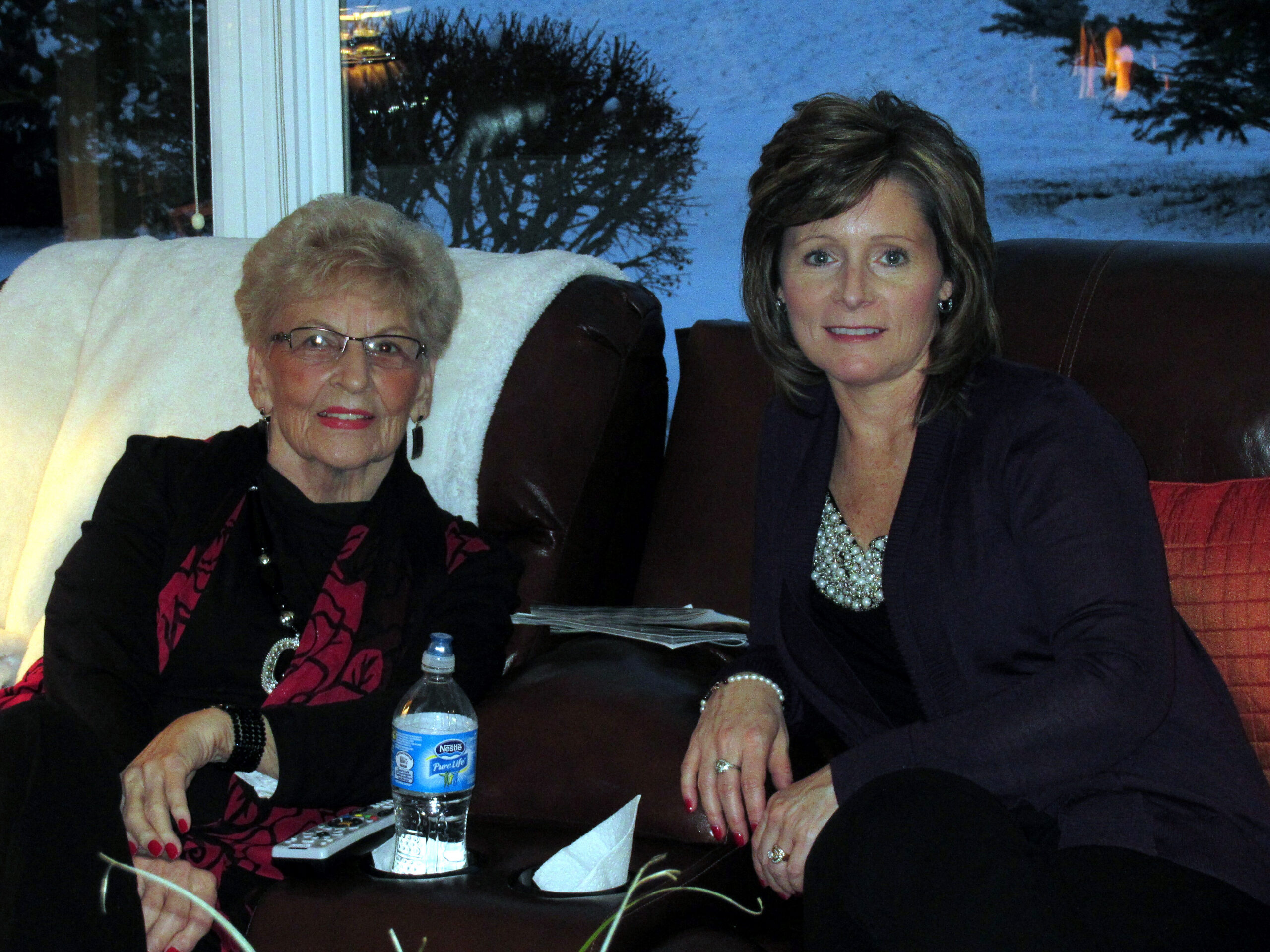Two women sit on a brown leather couch in a living room, smiling at the camera with a water bottle and remote on the table nearby.