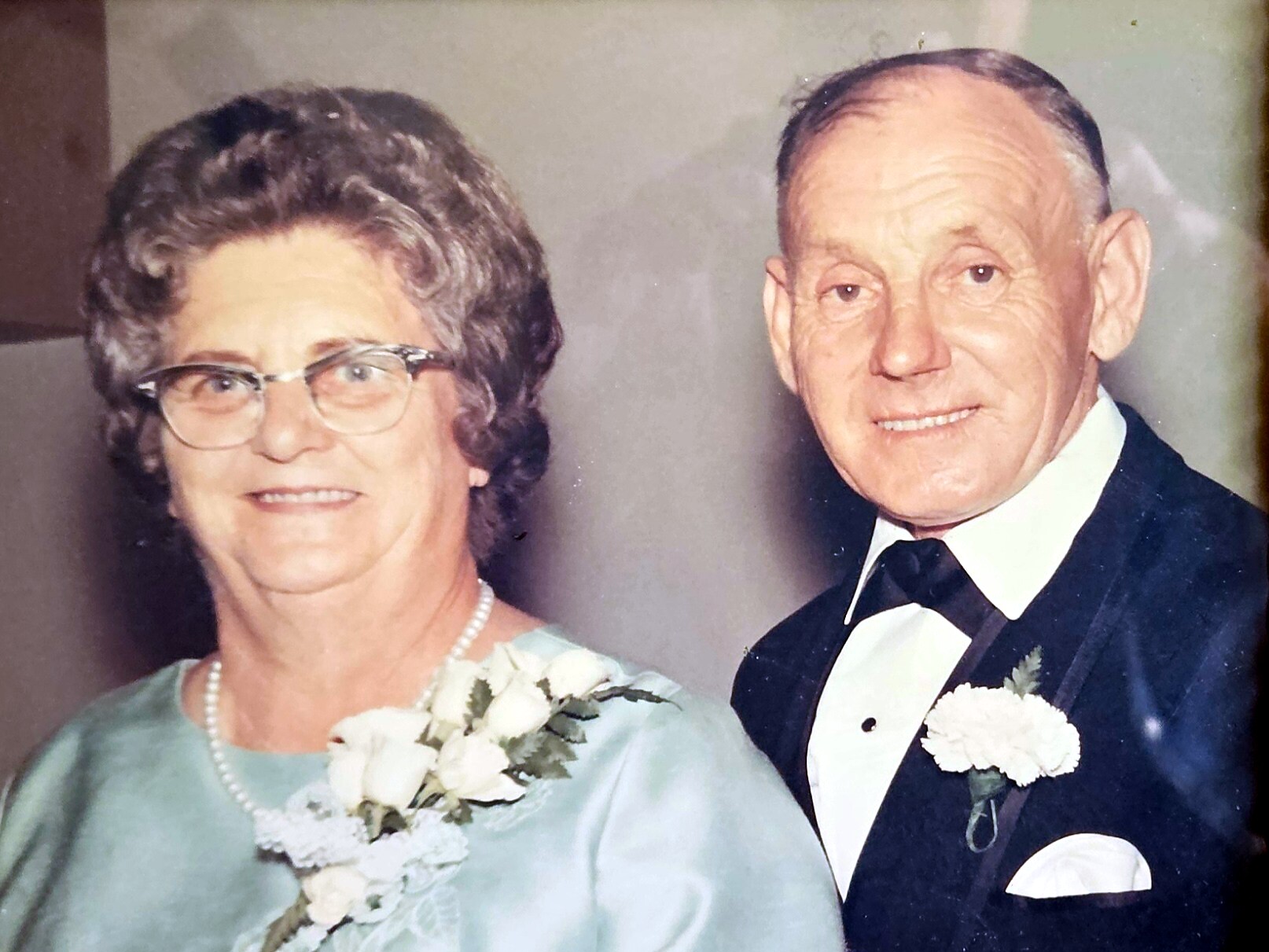 An elderly couple dressed for a formal event, smiling side by side in a portrait-style photo. The woman wears a light blue dress with a corsage and pearl necklace, the man wears a black tuxedo with a white boutonniere.