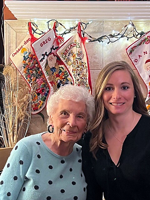 Two women posing indoors with colorful Christmas stockings hanging behind them on a mantelpiece.