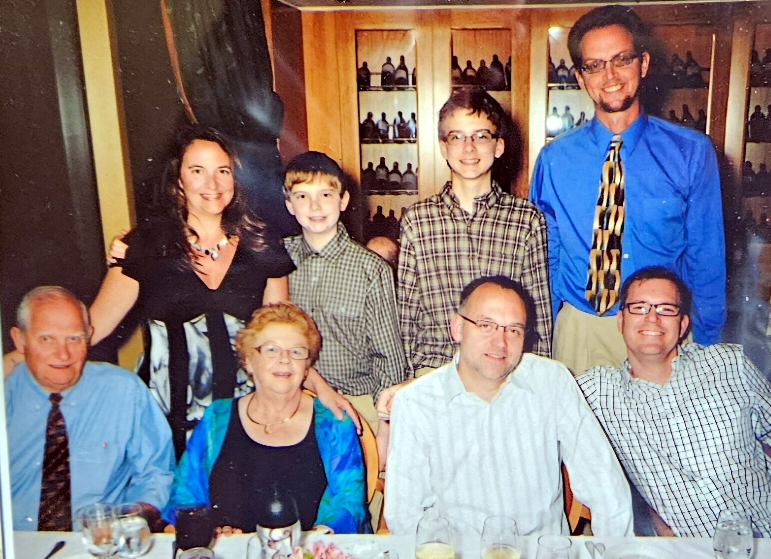 Group of people smiling for a photo at a social gathering, standing and seated around a table with drinks; warm, casual party setting in the background.