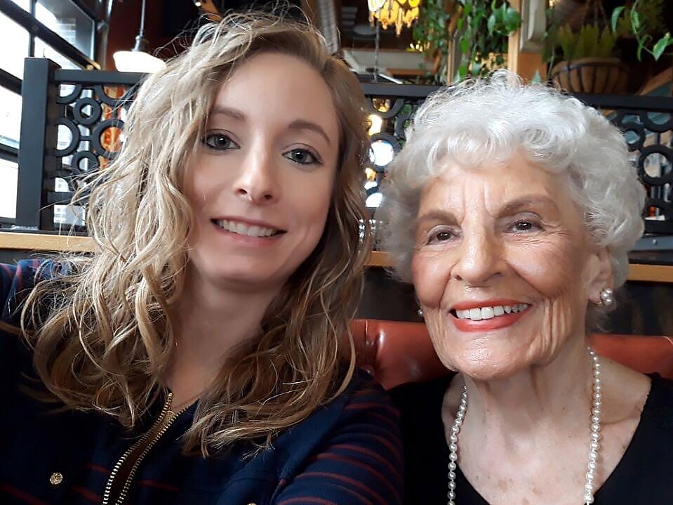 Two women smiling for a selfie in a cafe: a young woman with wavy blond hair and an elderly woman with white hair wearing pearls.
