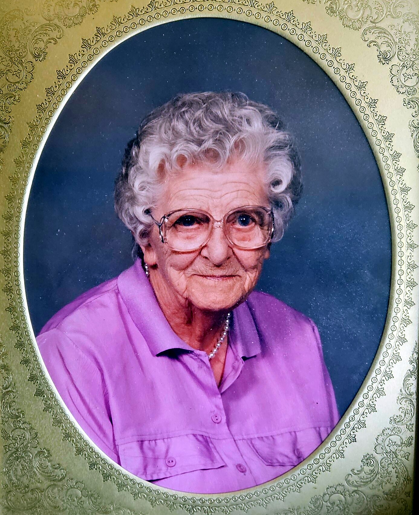 Portrait of an elderly woman with short curly white hair and glasses, wearing a purple shirt and pearl necklace, in a decorative gold frame.