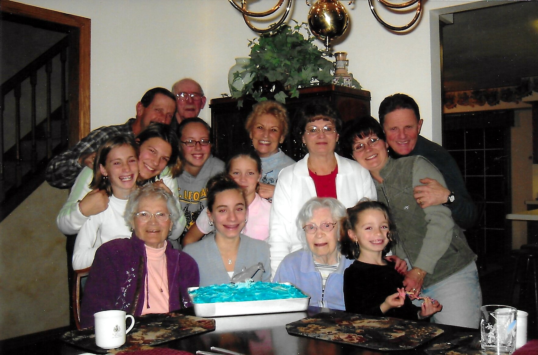 A large group of family members smiling and posing together around a dining table with a blue‑frosted cake in front, indoors by a staircase and decorative china cabinets in the background.