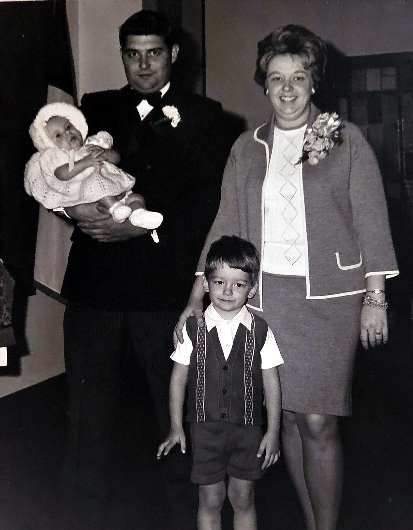 Formal family portrait: man in a suit holding a baby in a white crochet outfit, woman in a patterned dress with a corsage, and a young boy in a vest standing in front.