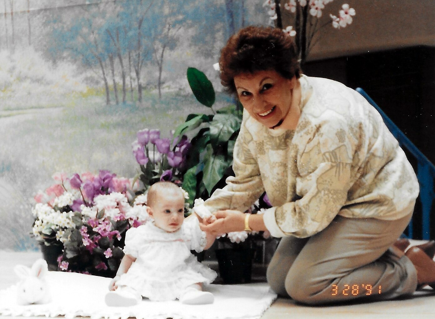 Woman kneeling beside a baby in a white dress, holding the baby's hand as they sit on a white mat near colorful flowers and a stuffed bunny.