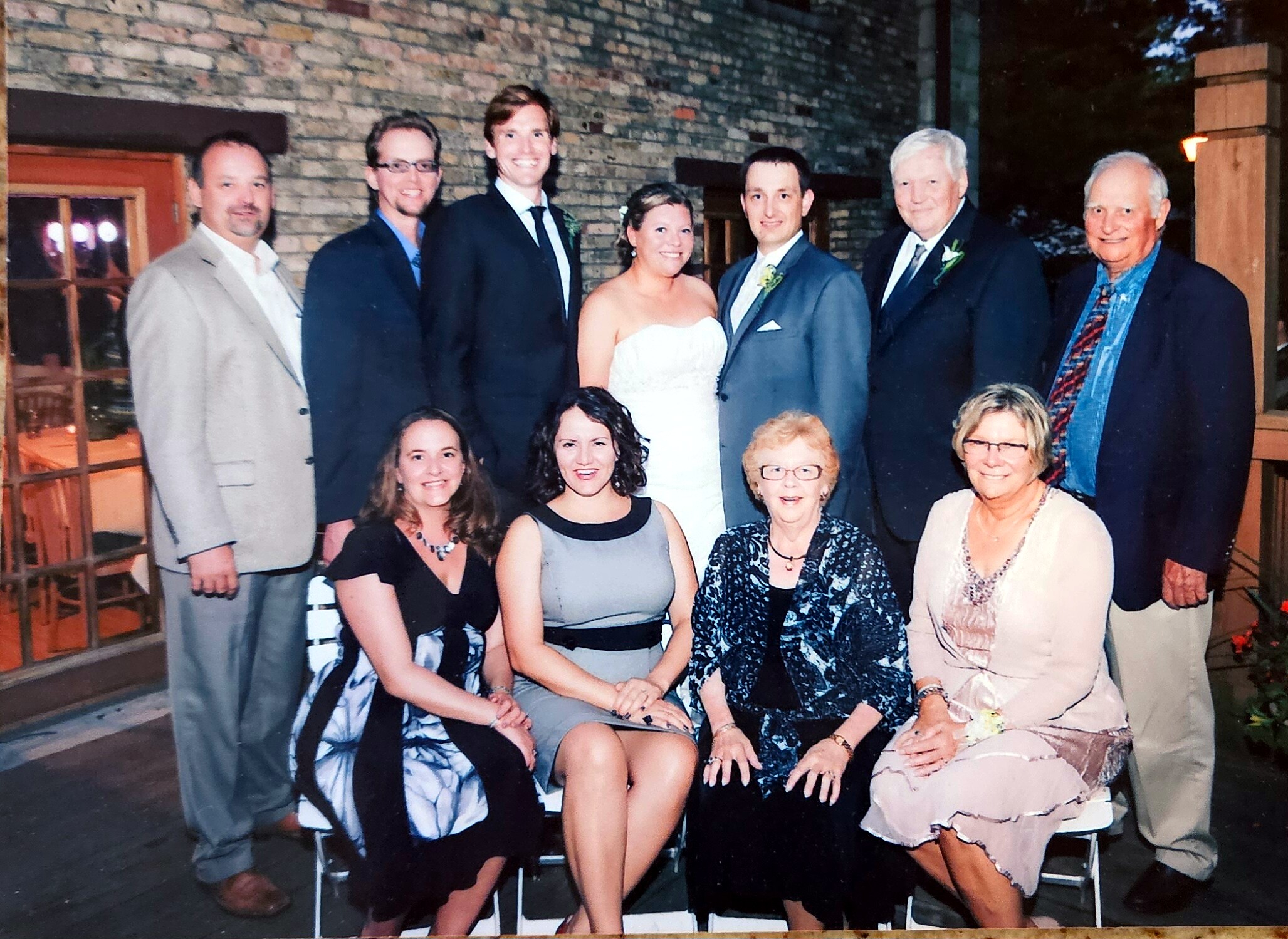 Group wedding party posing outside a building: bride in a white strapless gown and groom in a gray suit stand center, flanked by friends and family.