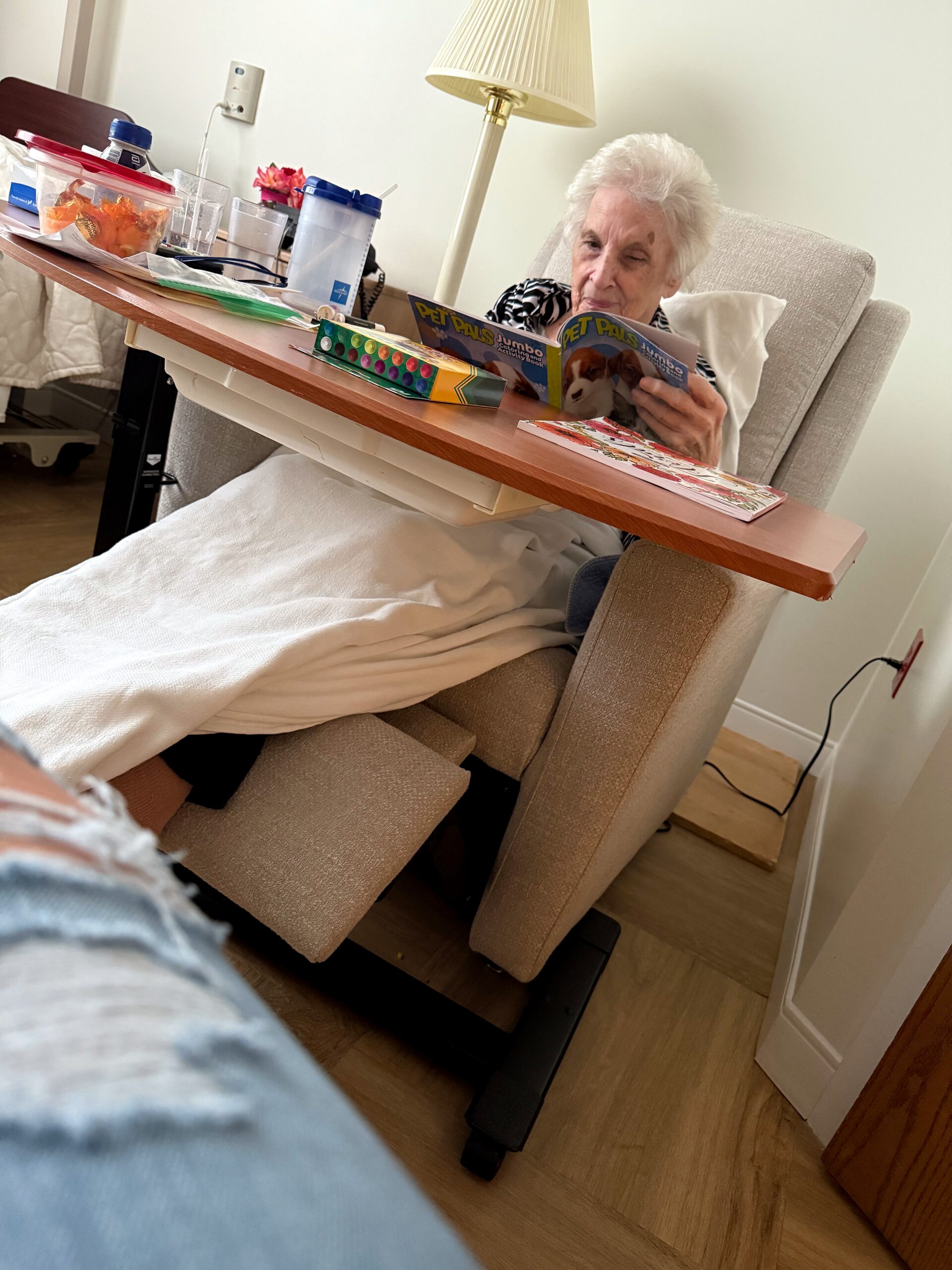 Elderly woman in a beige chair reads a 'Pet Pals' magazine on a raised bed tray with art supplies nearby.