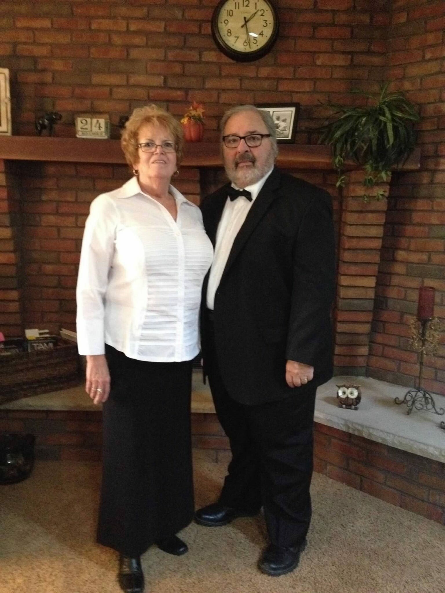 Older couple posing indoors in front of a brick fireplace; man in a tuxedo and bow tie, woman in a white blouse and black skirt.
