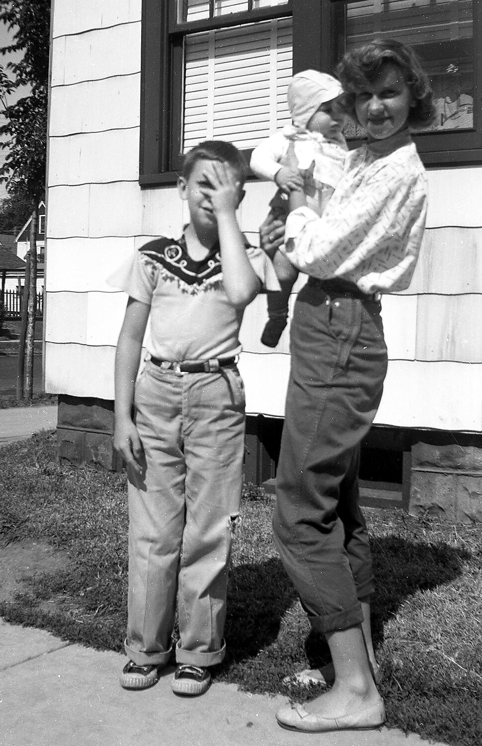 A woman holding a baby stands beside a boy outside a house; the boy covers his face with his hand.
