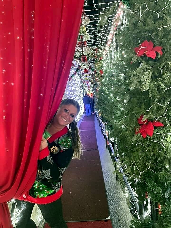 Smiling woman peeking from behind a red curtain into a festive, light-filled Christmas tunnel with greenery and poinsettias behind her.