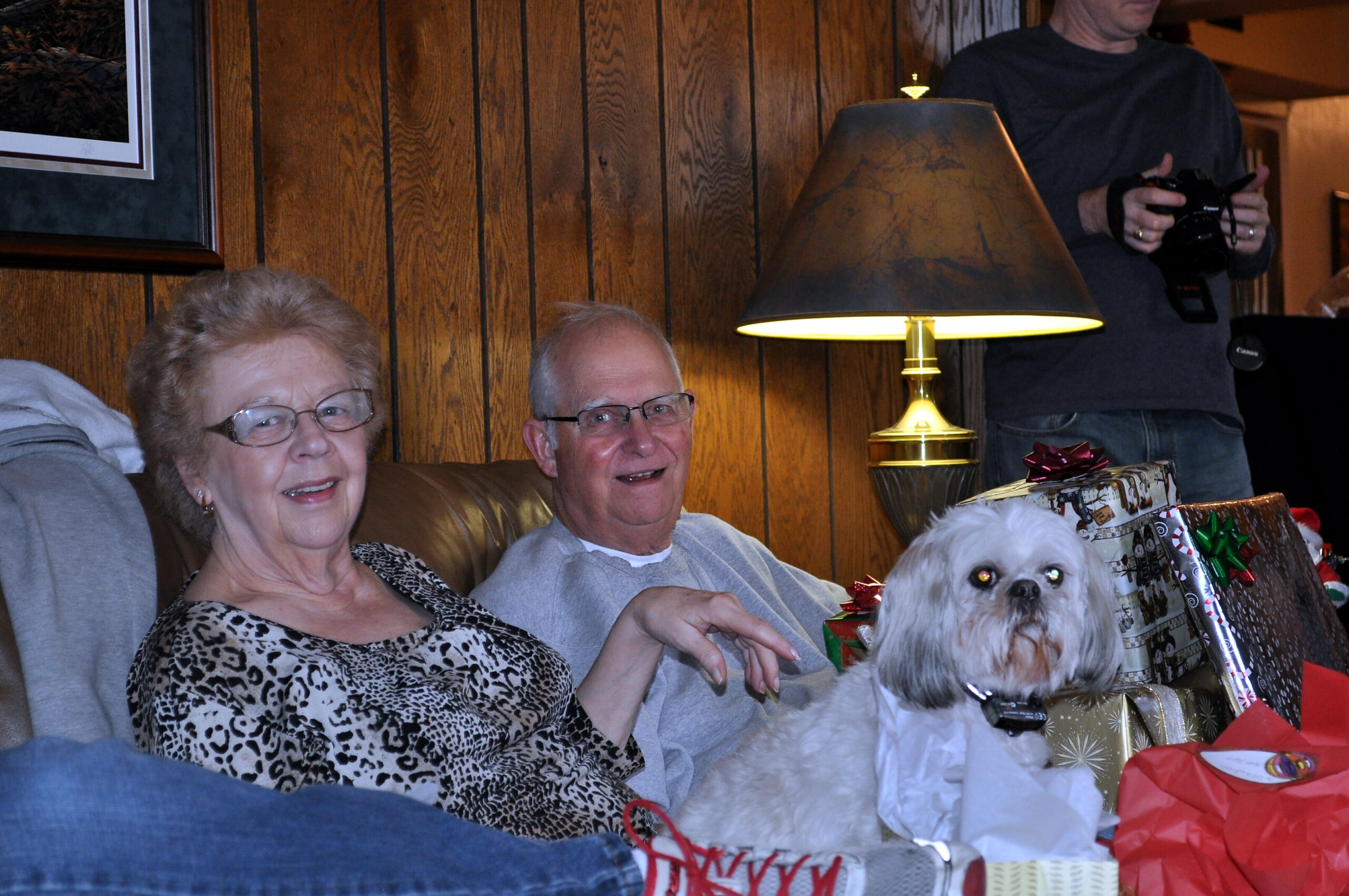 Smiling elderly couple on a sofa with a small fluffy dog and wrapped gifts in a cozy wood-paneled living room.