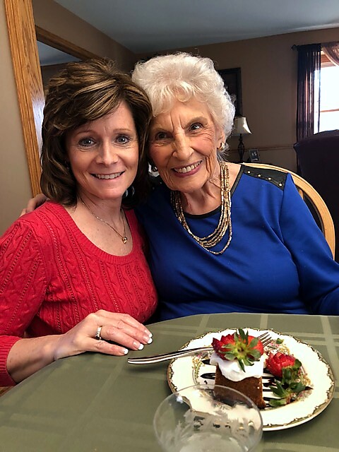 Two women smiling at a dining table, with a chocolate cake dessert topped with berries in the foreground.
