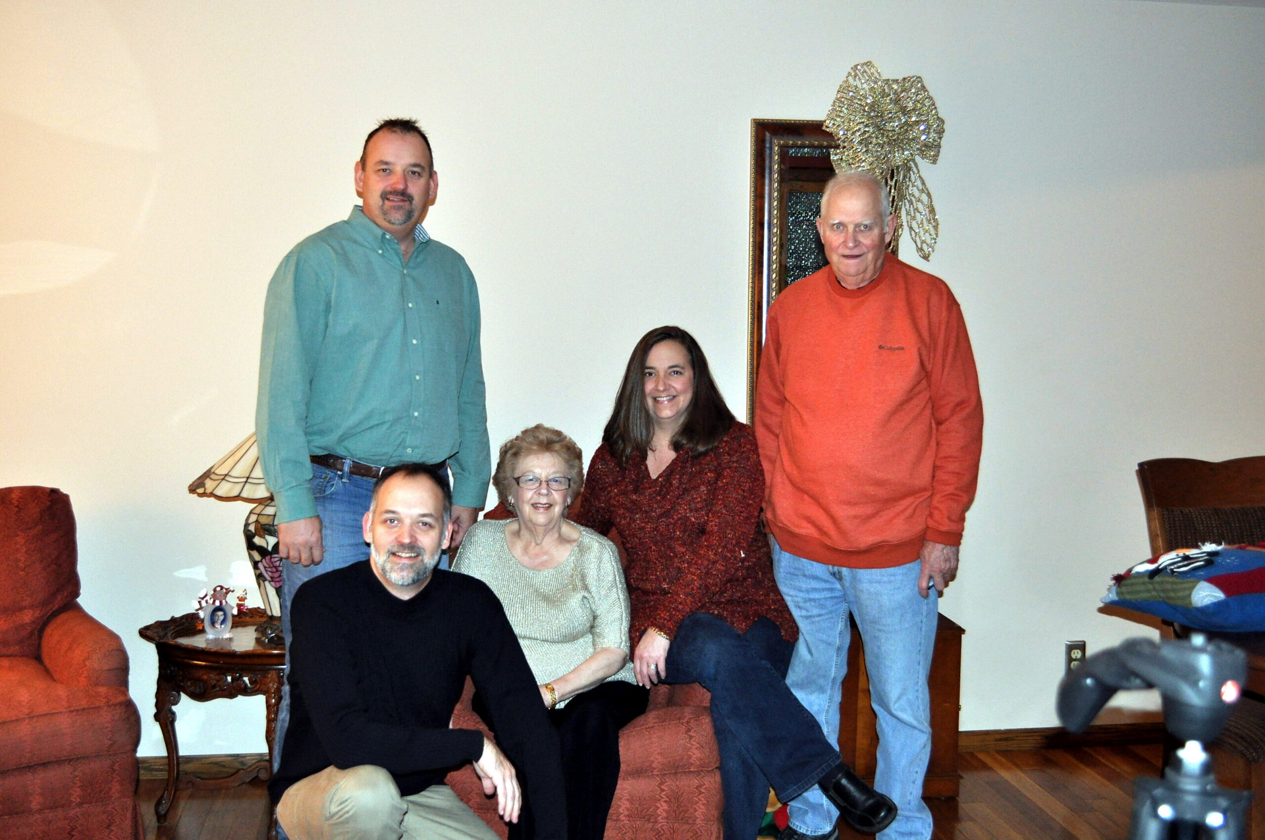 Group of six people posing for a family photo in a living room with red furniture and holiday decor in the background, including a large silver bow on a mirror.