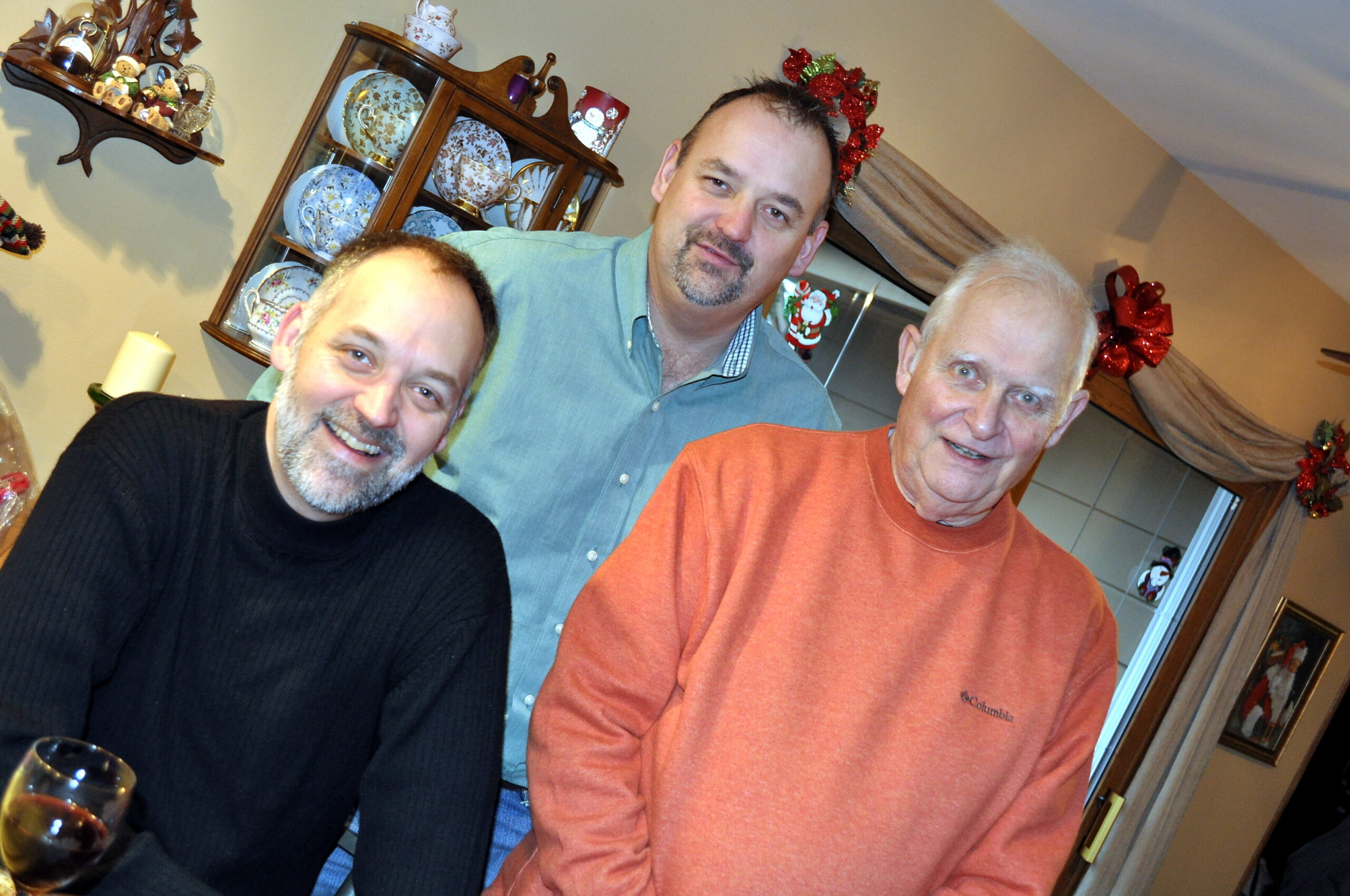 Three men smiling for a casual indoor photo at a Christmas gathering, with a china cabinet and holiday decor in the background.