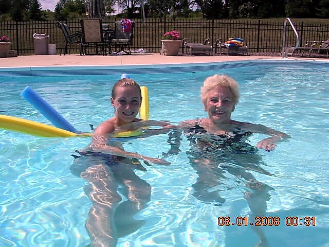 Two people in a bright backyard pool with foam pool noodles, smiling at the camera, patio furniture and fence in the background.
