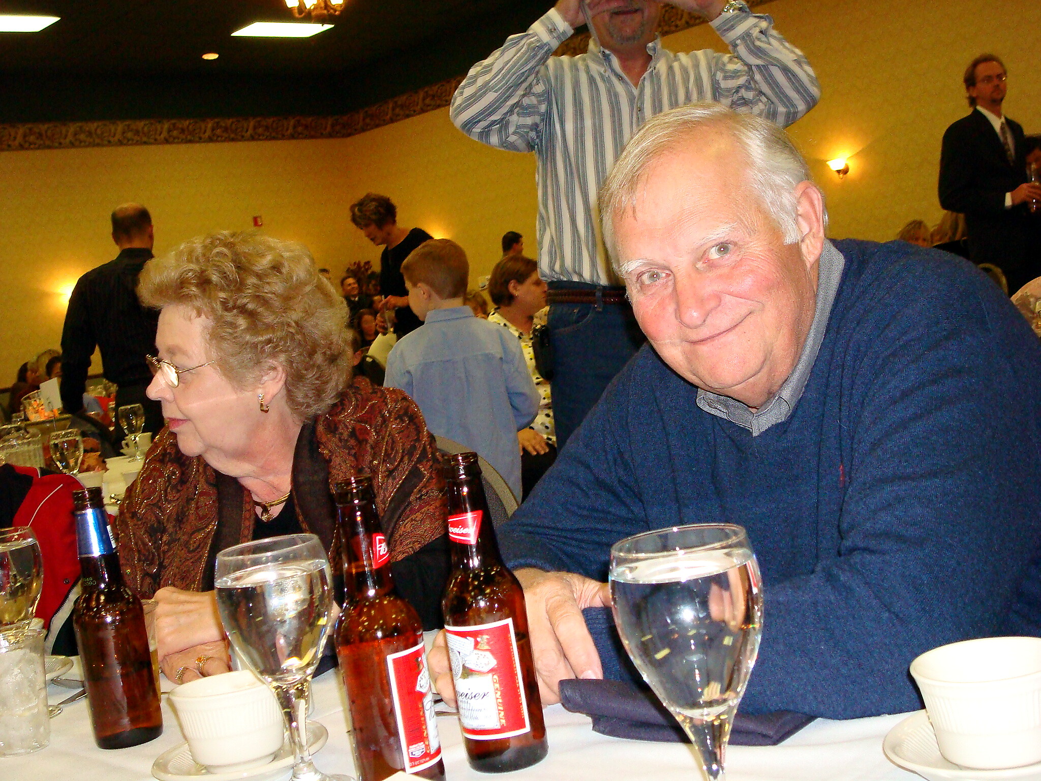 Older man in a blue sweater leans toward the camera at a crowded banquet table with beer bottles, wine glasses, and place settings in the foreground.