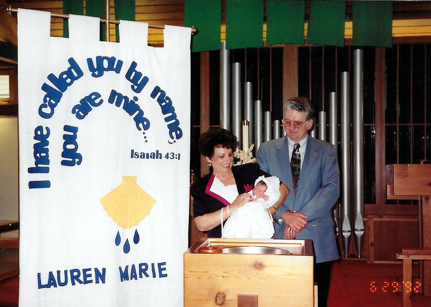 Woman with a baby in a white christening gown beside a man in a suit at a baptism ceremony; banner reads Isaiah 43:1 and the name Lauren Marie.