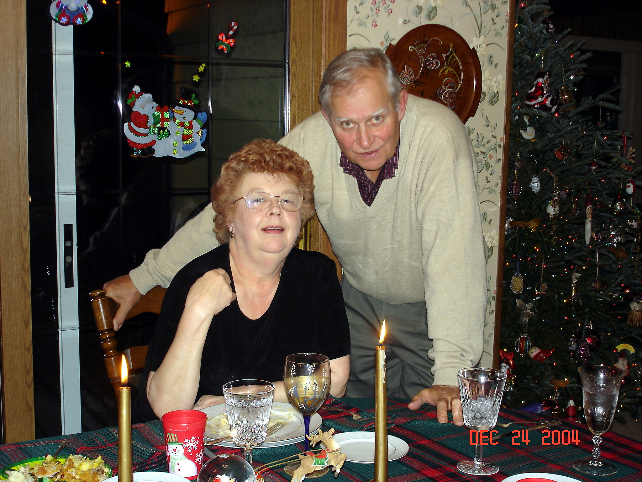 An elderly couple posing at a Christmas dinner, with candles on the table and a decorated Christmas tree in the background.