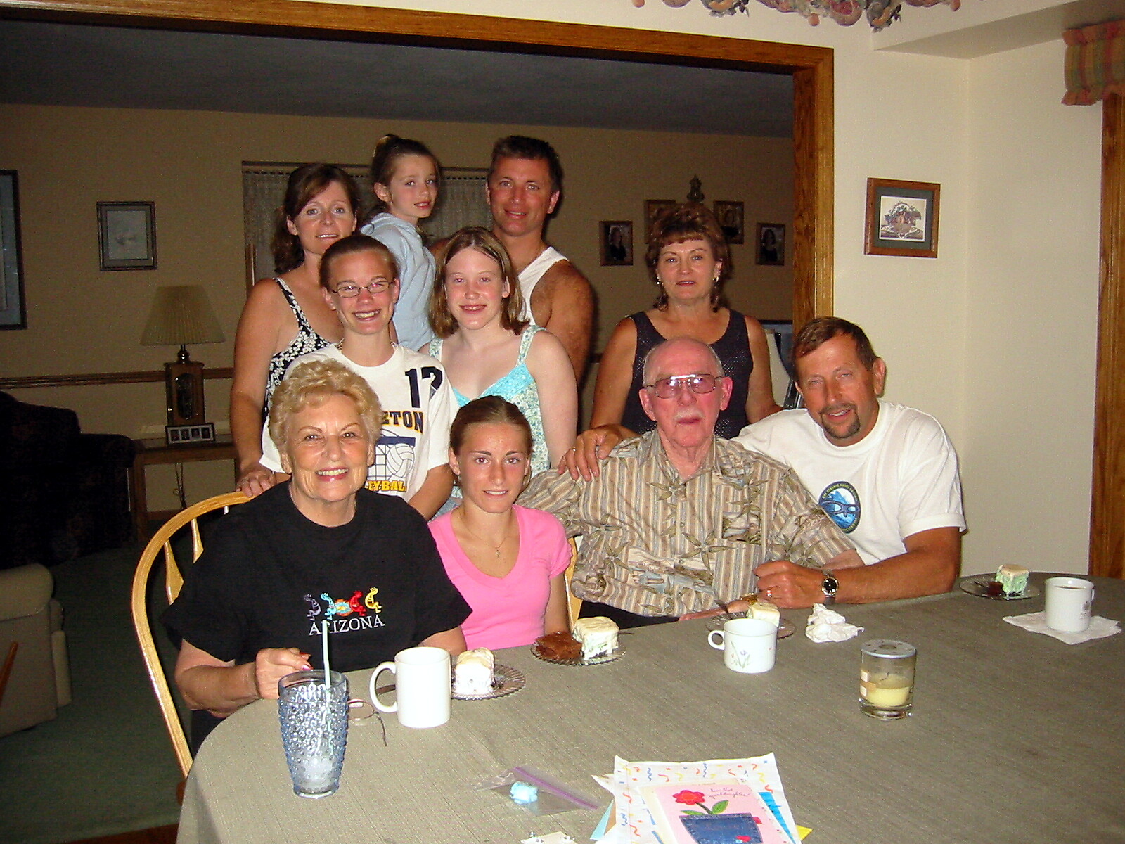 Group of family members of various ages gathered around a dining table with cupcakes and drinks, posing for a photo.