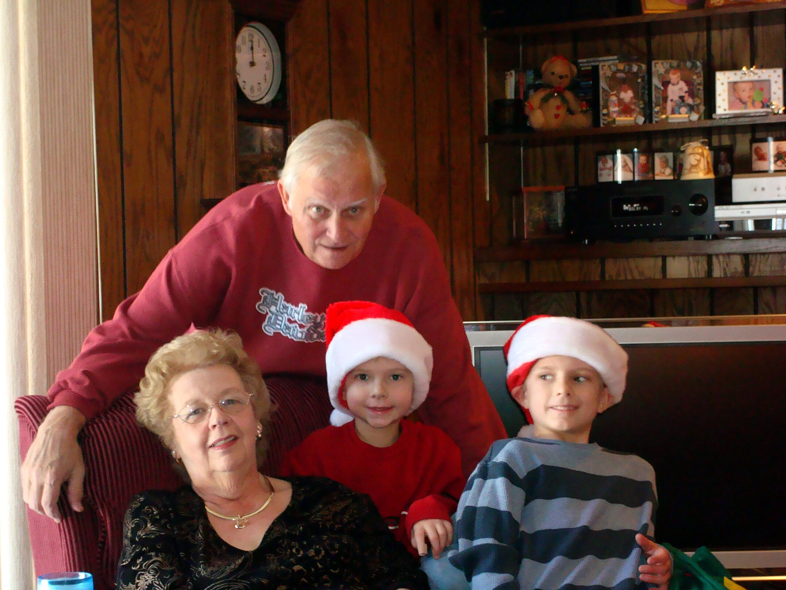 Family group posing indoors for a holiday photo, with two adults and three children wearing Santa hats on a couch.