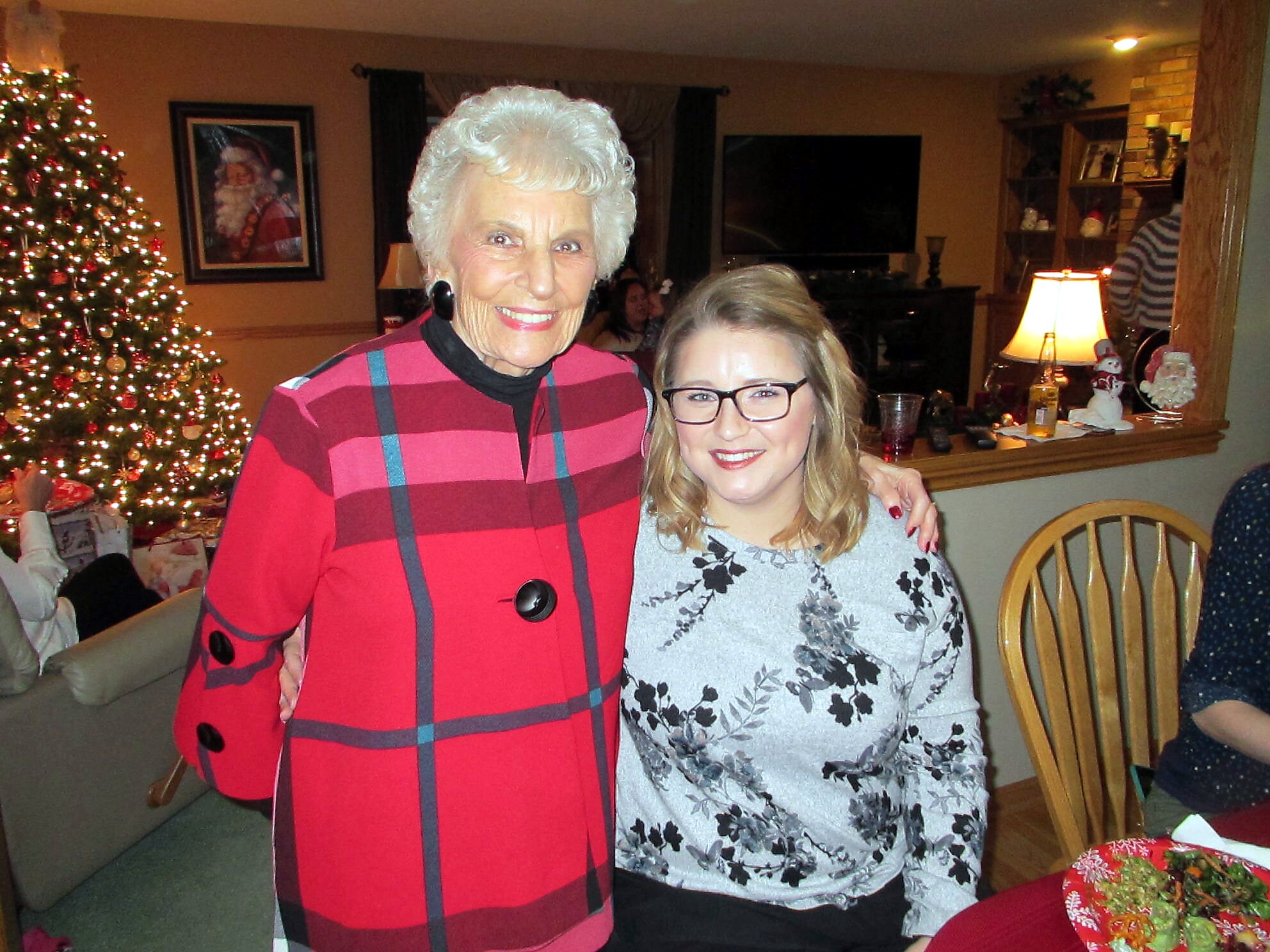Two smiling women pose together in a festive living room with a Christmas tree in the background; one wears a red plaid coat and the other a floral sweater.