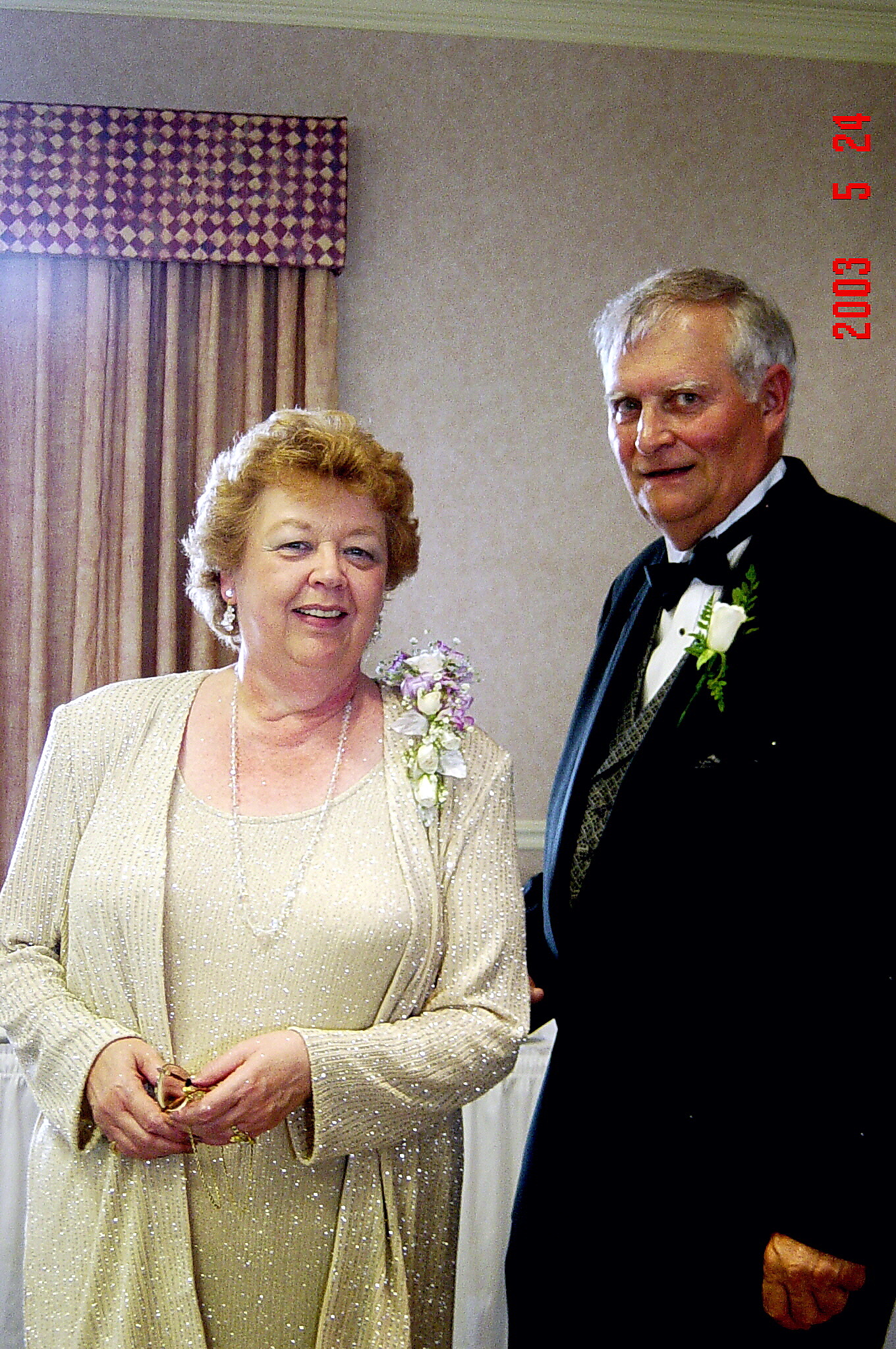 Smiling elderly couple at a formal event: a woman in a champagne sequined dress and shawl, and a man in a black tuxedo with a boutonniere, posing indoors.