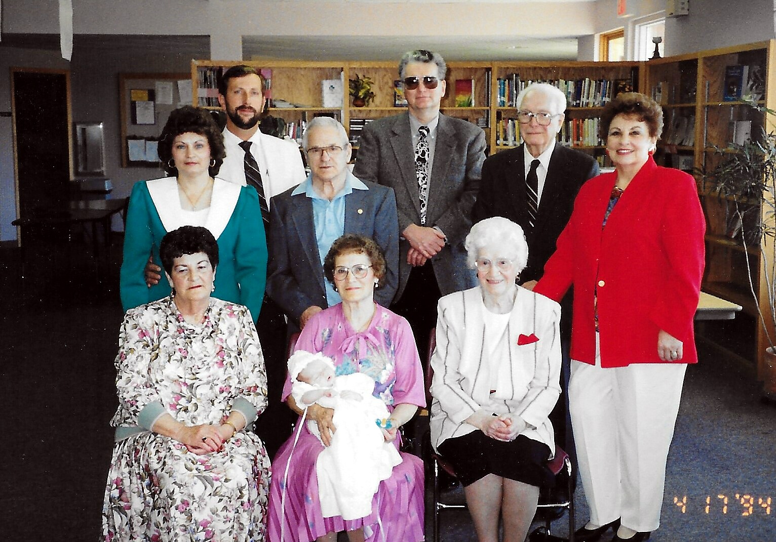 Group of nine adults gathered for a portrait in a library, with bookshelves in the background; two women seated in front, one holding a baby wrapped in white, others stand behind them in colorful outfits.
