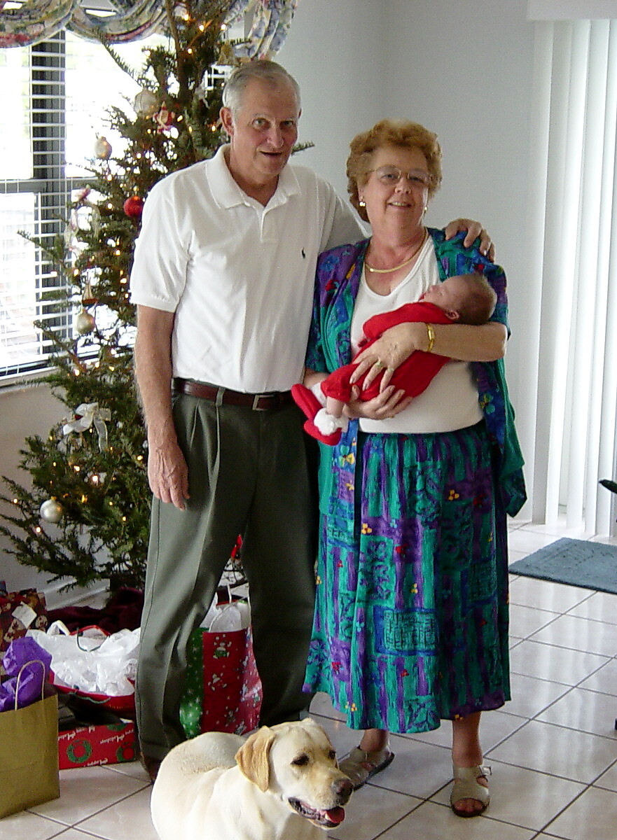 Grandfather and grandmother stand together holding a newborn baby in a living room with a Christmas tree and wrapped gifts nearby, a golden retriever sits in front.