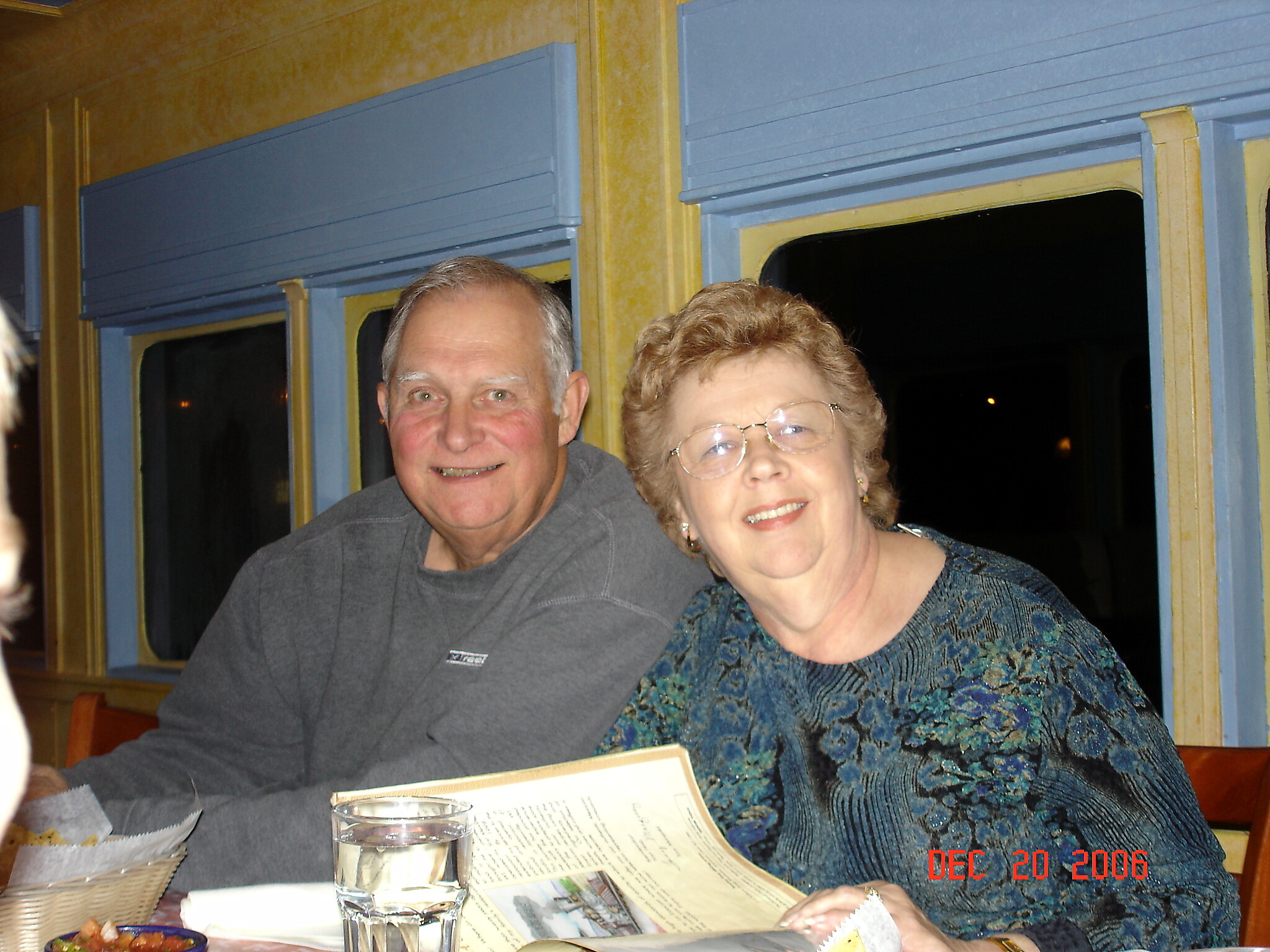 Smiling older man and woman seated together at a dining-table on a train, with a newspaper and a glass of water in front of them.