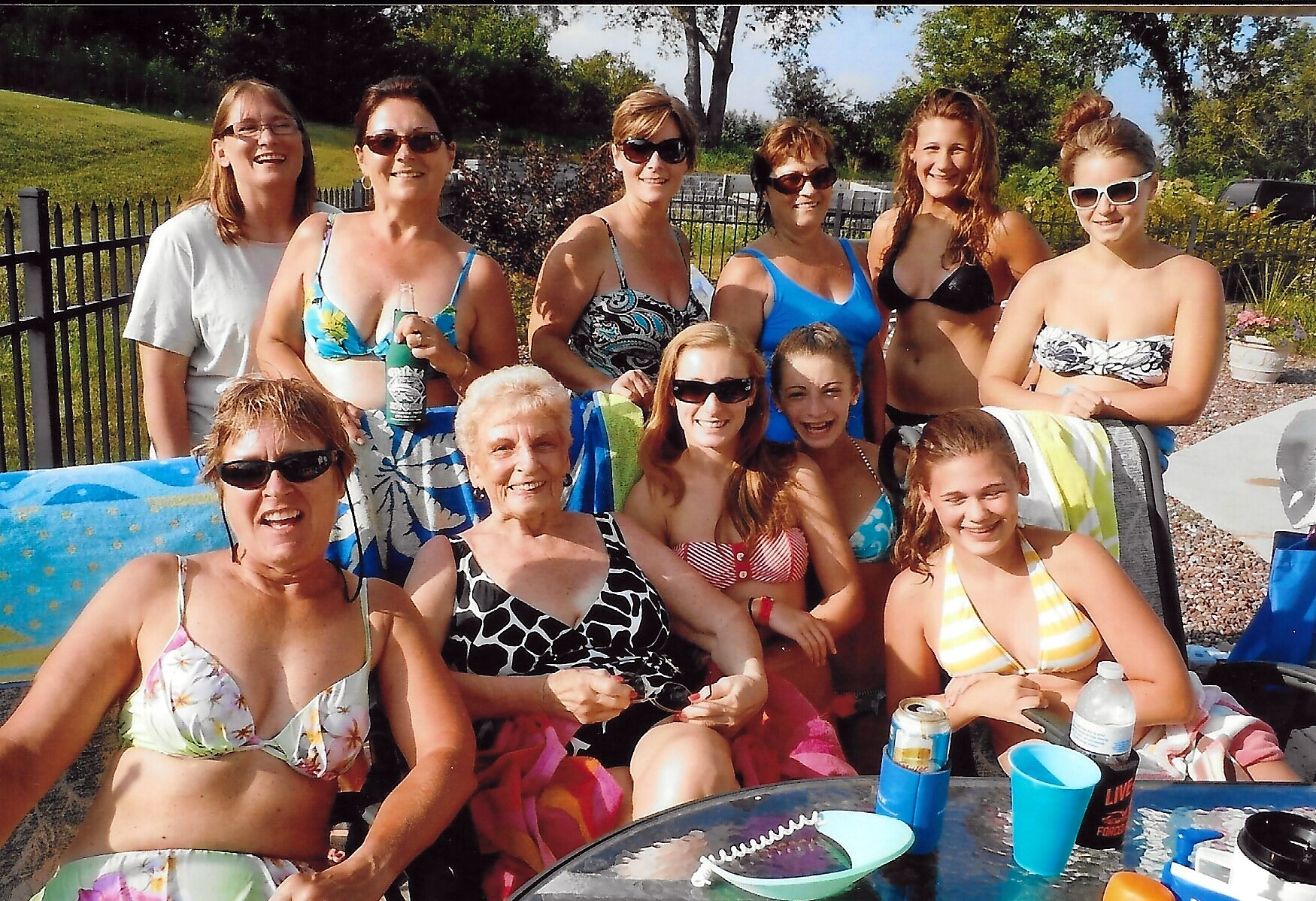 Group of women in swimsuits posing and smiling poolside at an outdoor gathering, with towels and drinks nearby.