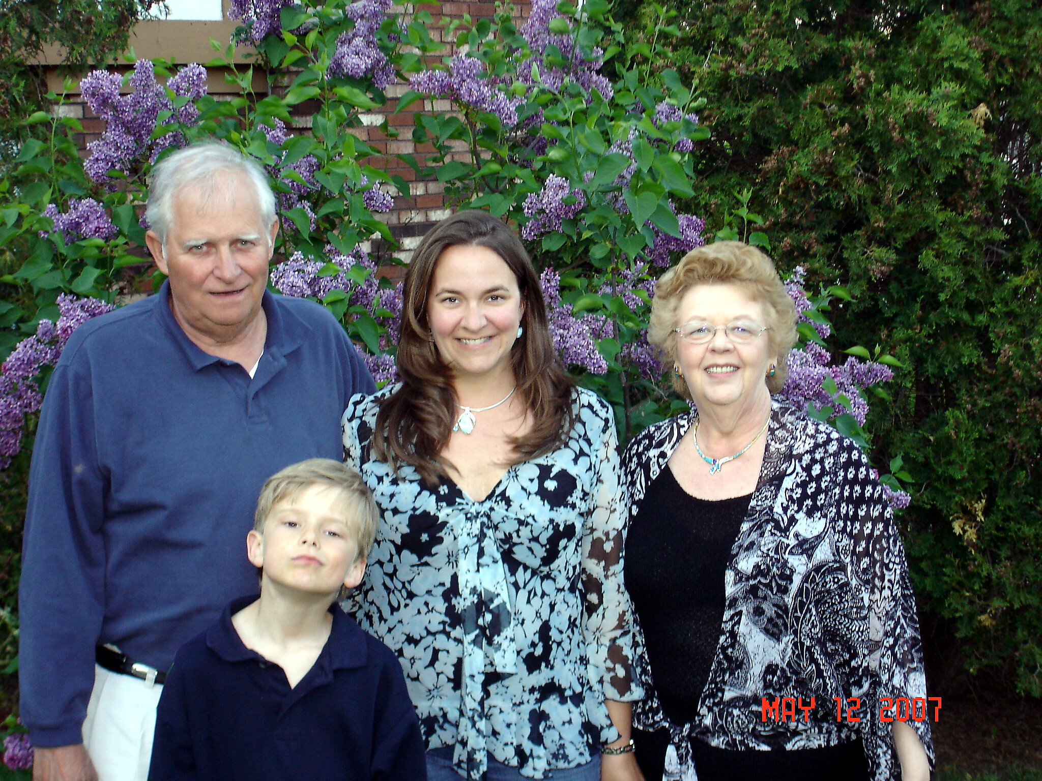 Family group posing outdoors in front of lilac bushes and a brick wall, with a smiling man on the left, a woman in the center, an older woman on the right, and a boy in the foreground.