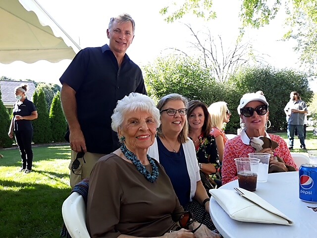 Group of smiling adults seated around a table at an outdoor garden party; elderly woman in front with turquoise necklace.