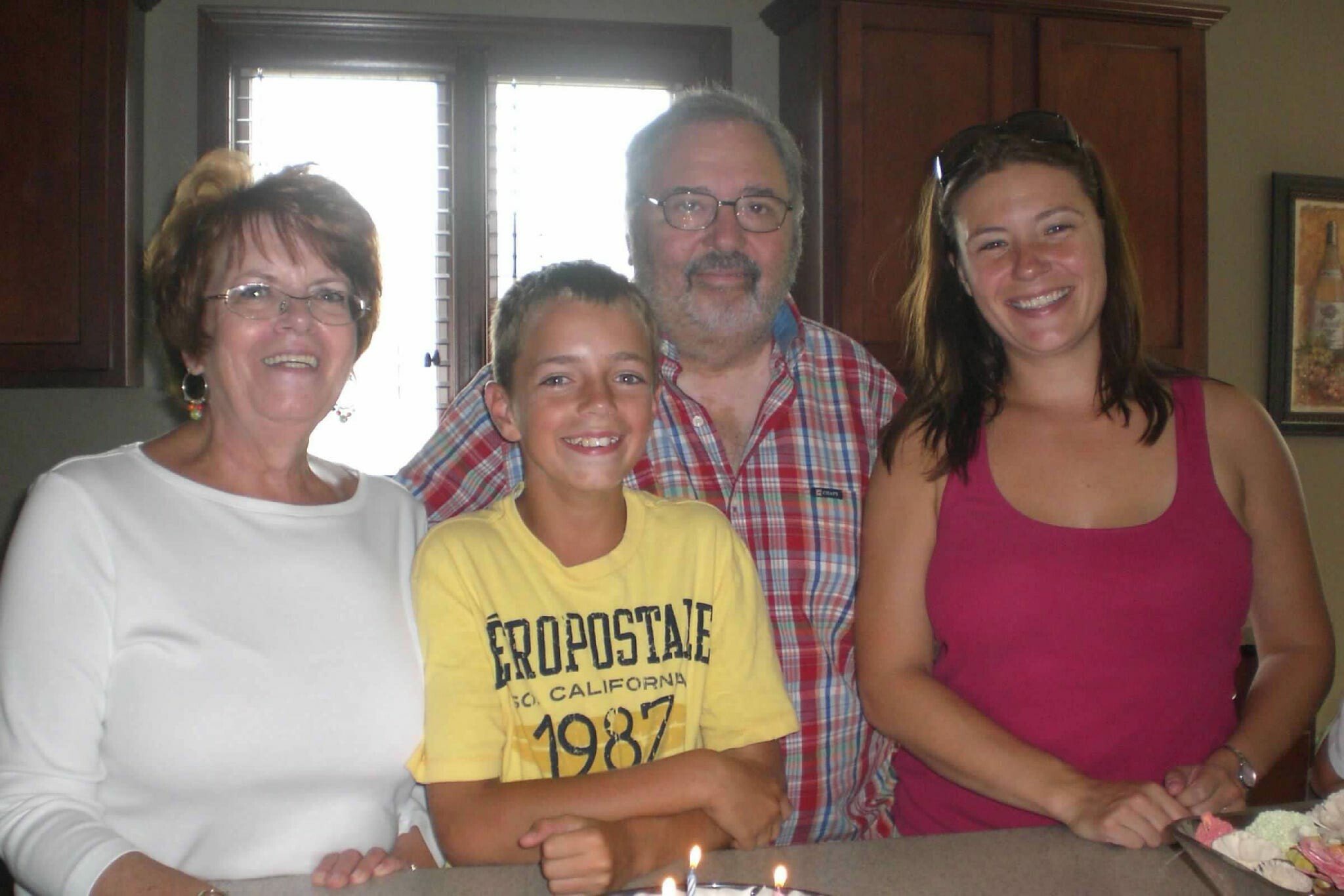 Family of four posing for a birthday photo in a kitchen, with a cake and lit candles on the counter in front of them.