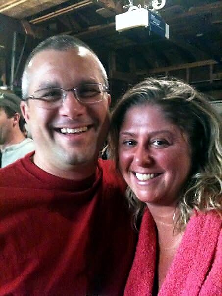 Two smiling adults posing for a photo at an indoor event, with a man wearing glasses in a red shirt and a woman in a red sweater nearby.