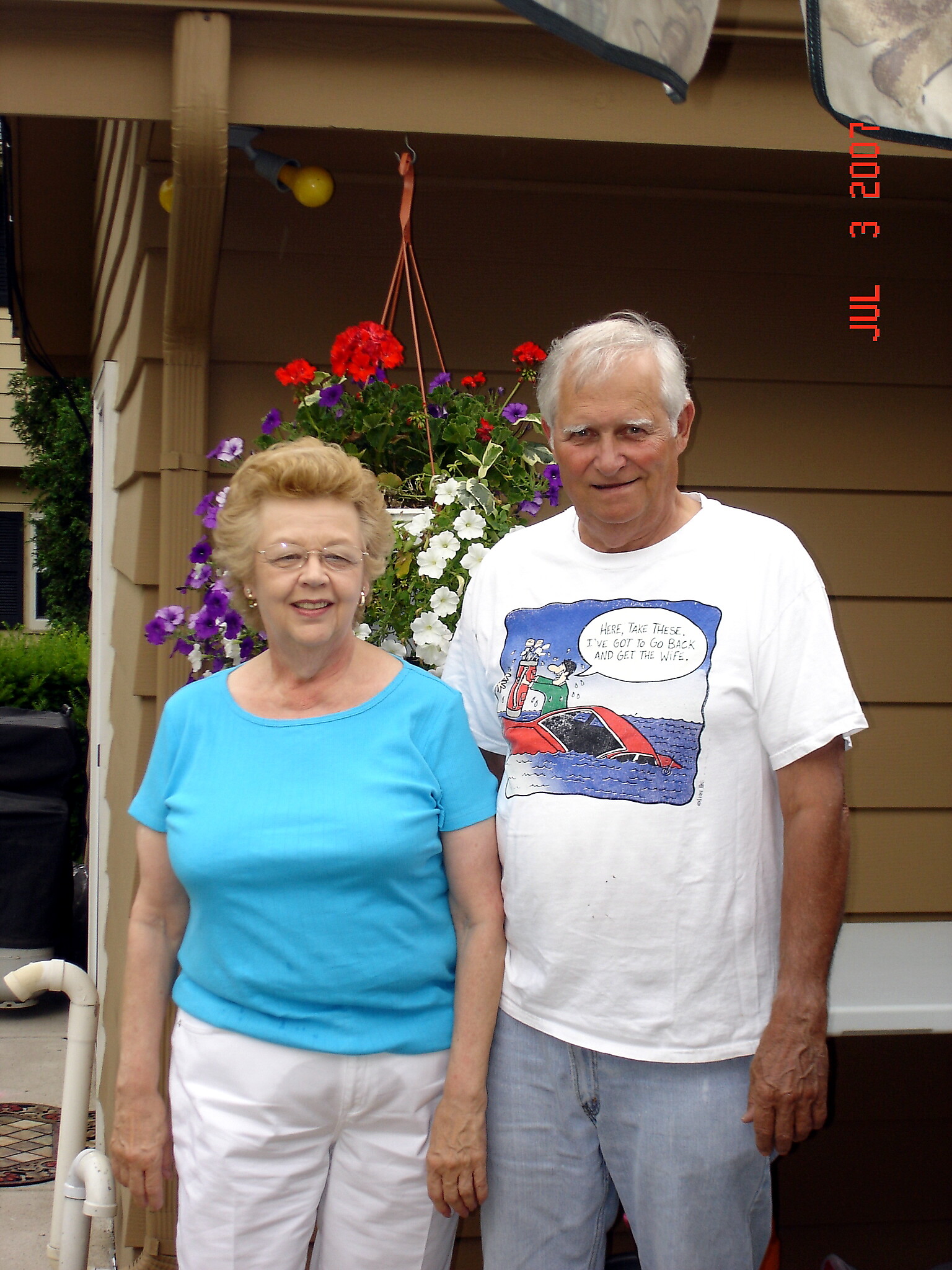 Smiling elderly couple standing outdoors in front of a house with a colorful hanging flower basket behind them.