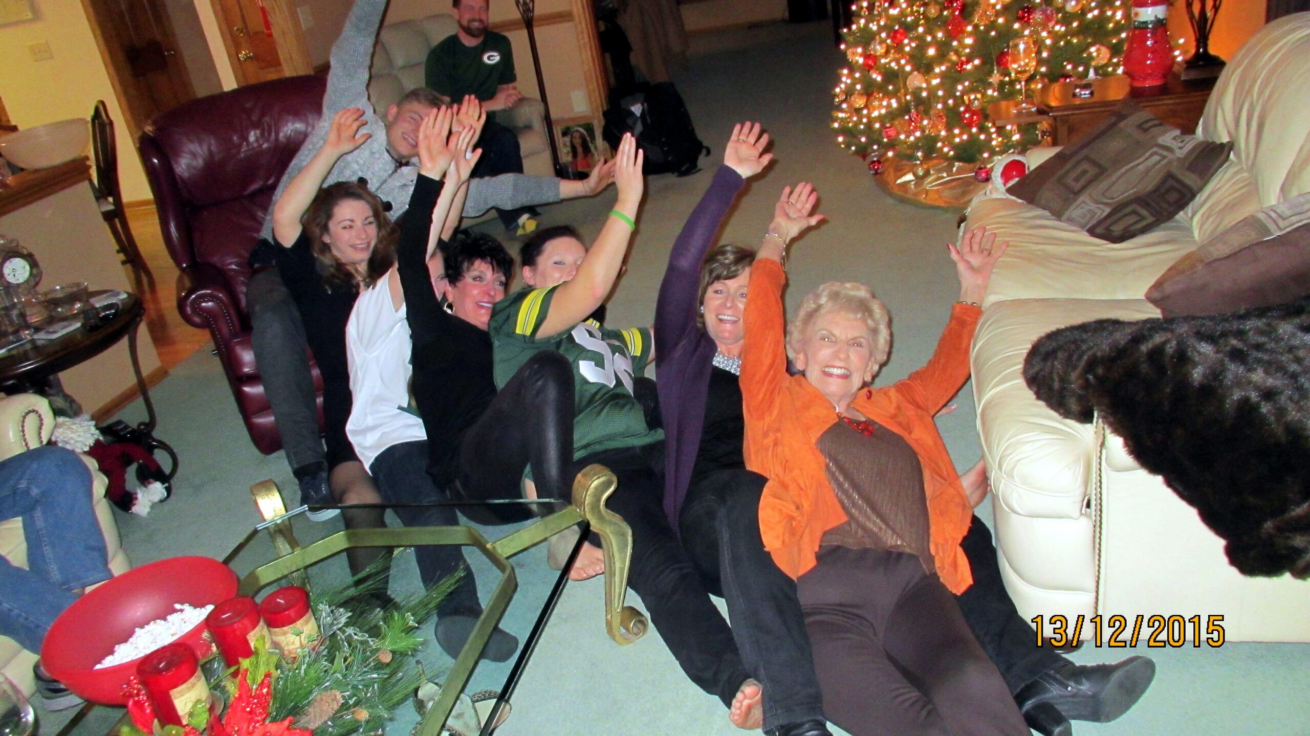 Group of adults on the living room floor with arms raised in a holiday party, Christmas tree with lights in the background.