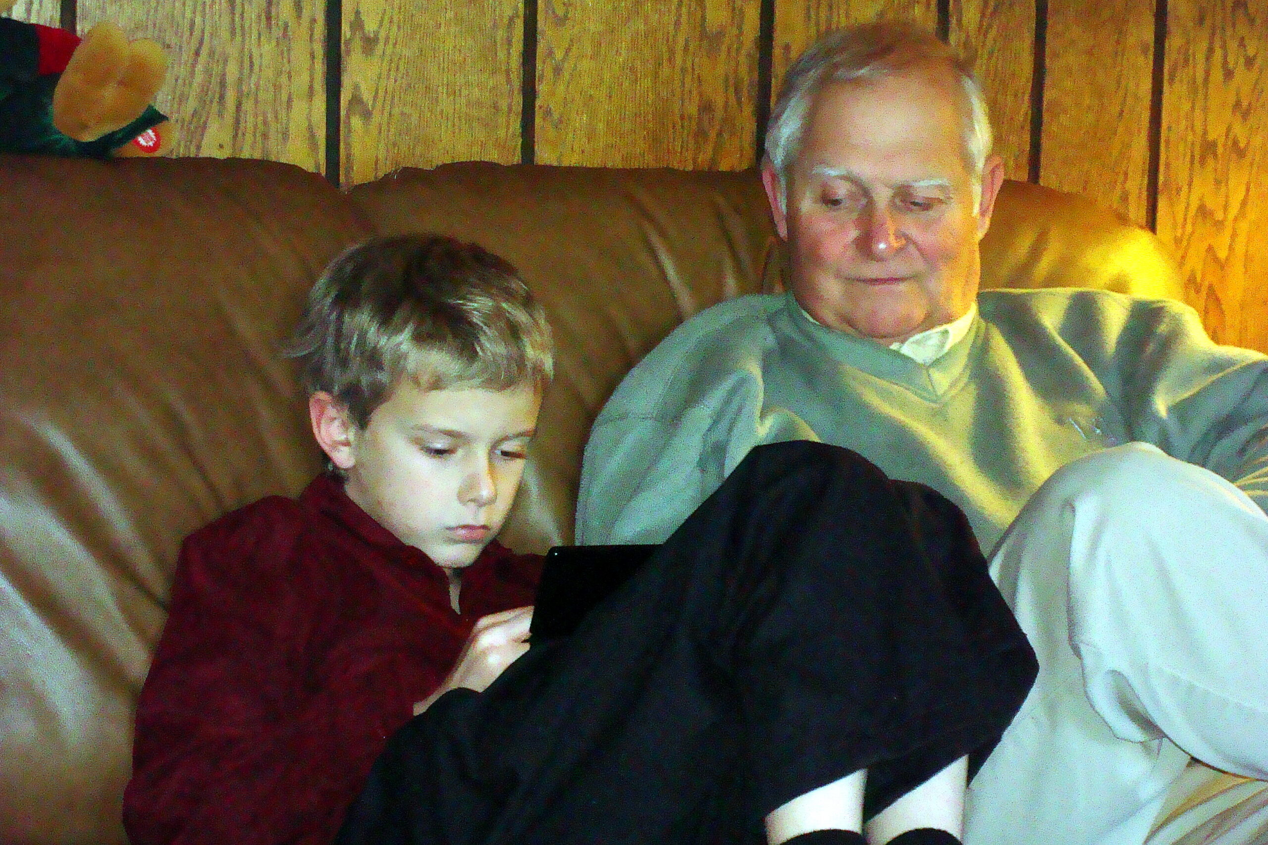 A young boy in a burgundy shirt sits beside an older man on a tan couch, both focused on a tablet device in the boy's hands/ lap.