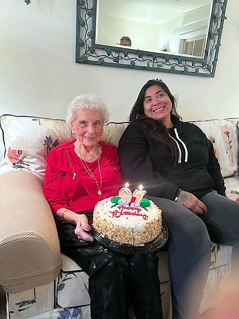 Elderly woman in red holds a birthday cake with lit 97 candles, seated on a floral sofa with a smiling companion beside her.