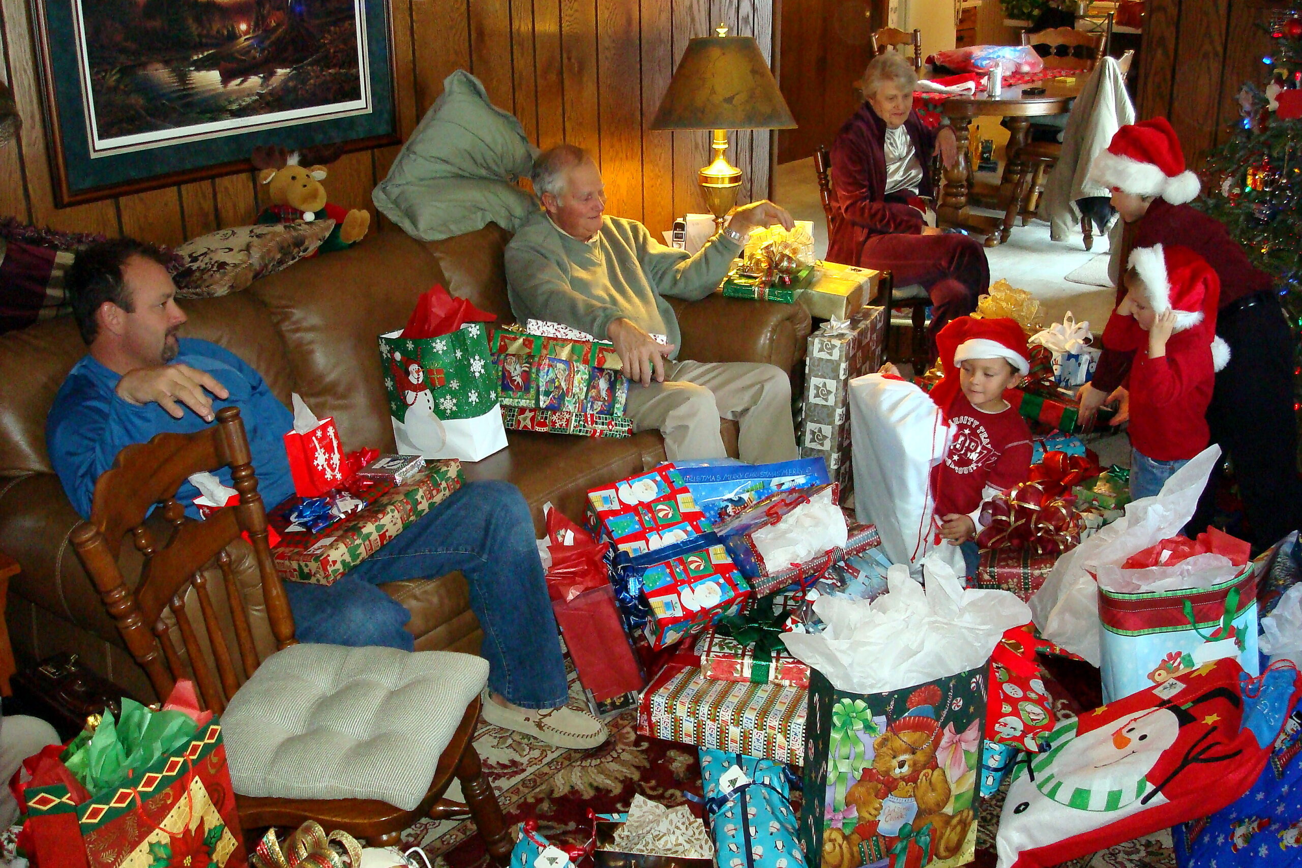 Living room full of wrapped Christmas presents around a man on a sofa as a child receives a gift; others wear Santa hats in a cozy, wood-paneled room.