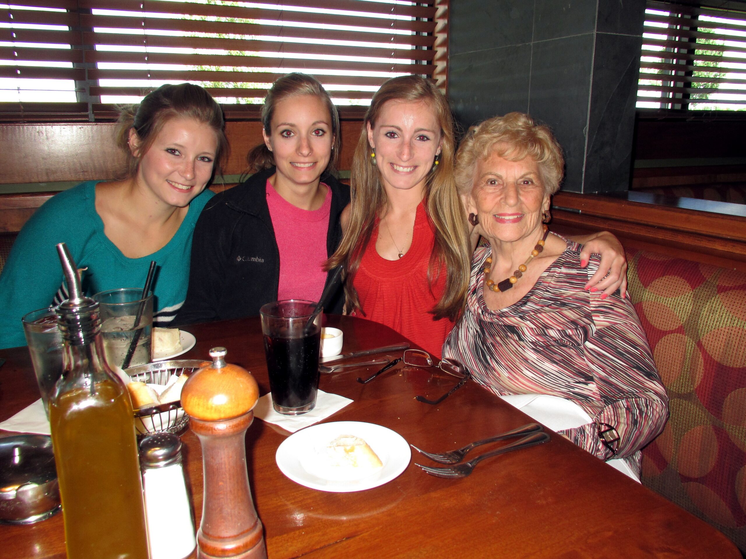 Four women pose together in a restaurant booth, smiling, with drinks and bread on the table in front of them.