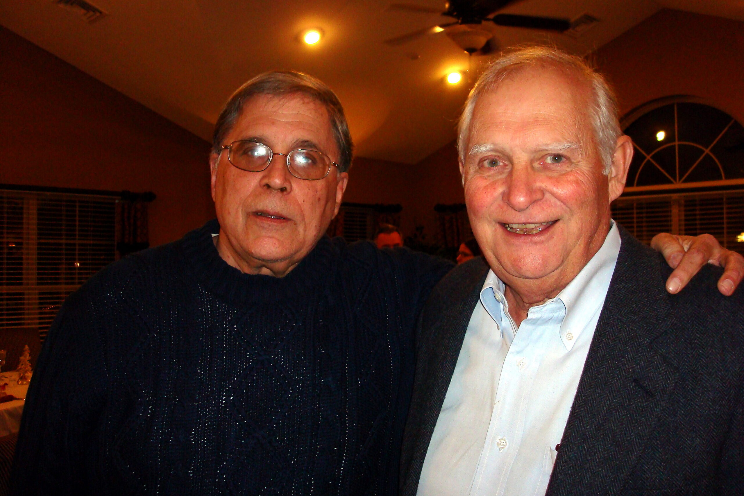 Two older men pose together indoors at a social gathering, smiling with their arms around each other to the camera.