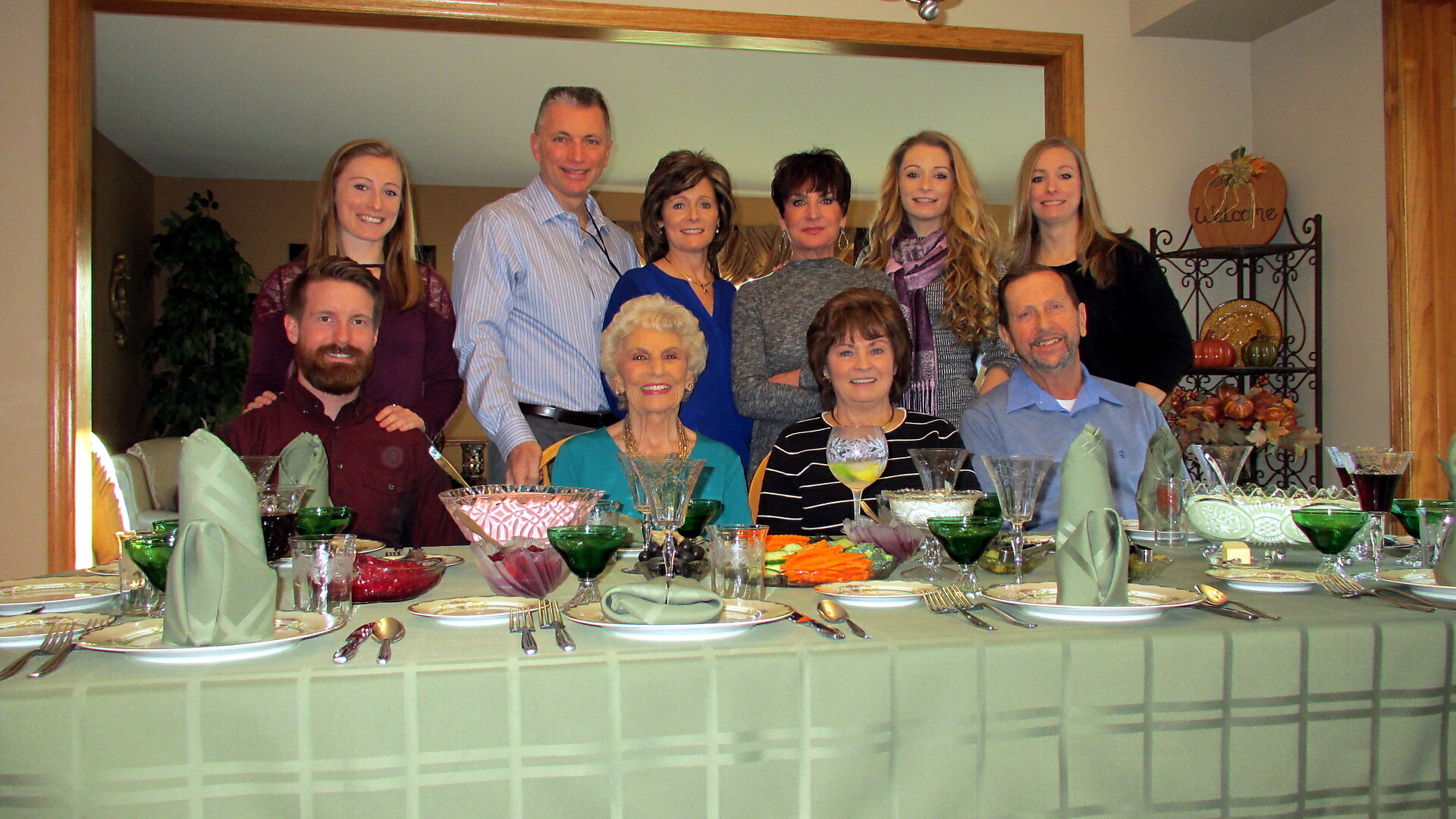 Group of adults posing for a group photo around a formally set dining table in a home, smiling at the camera.