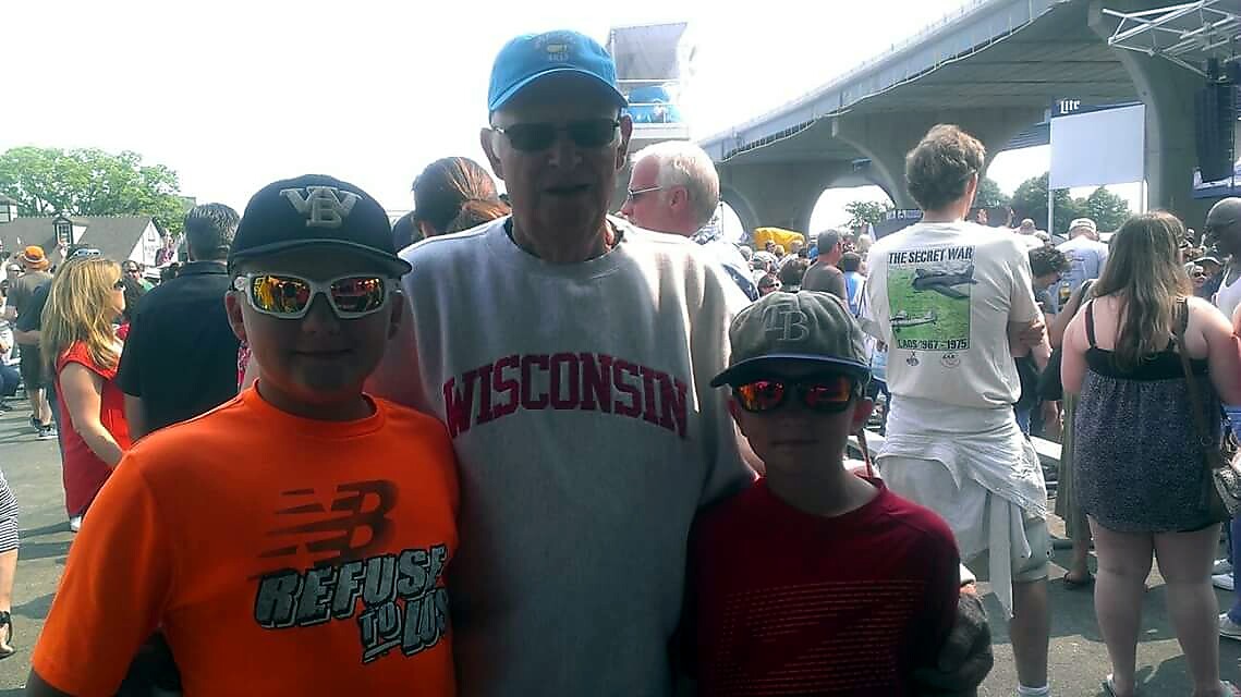 Three people pose together at a sunny outdoor festival; a man in a Wisconsin sweatshirt stands between two boys wearing caps and mirrored sunglasses with a crowd and stage in the background.