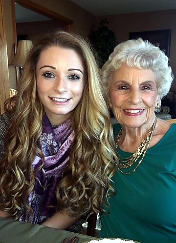 Young woman with long wavy blonde hair and a purple scarf sits beside an elderly woman with white hair; both smiling at the camera.