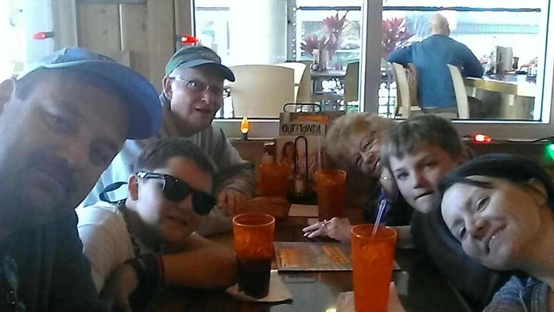 Six friends sit around a cafe table, taking a group selfie with orange drinks and straws visible on the table inside a bright dining area.
