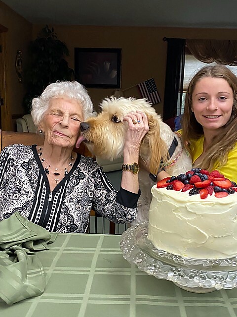 Old woman with white hair affectionately strokes a small beige dog at a birthday table, with a berry-topped cake and a smiling younger woman nearby.