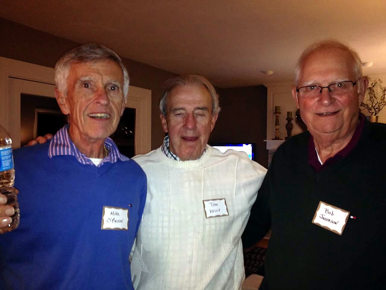 Three older men posing together at a party; the left man holds a bottle and all wear name tags.