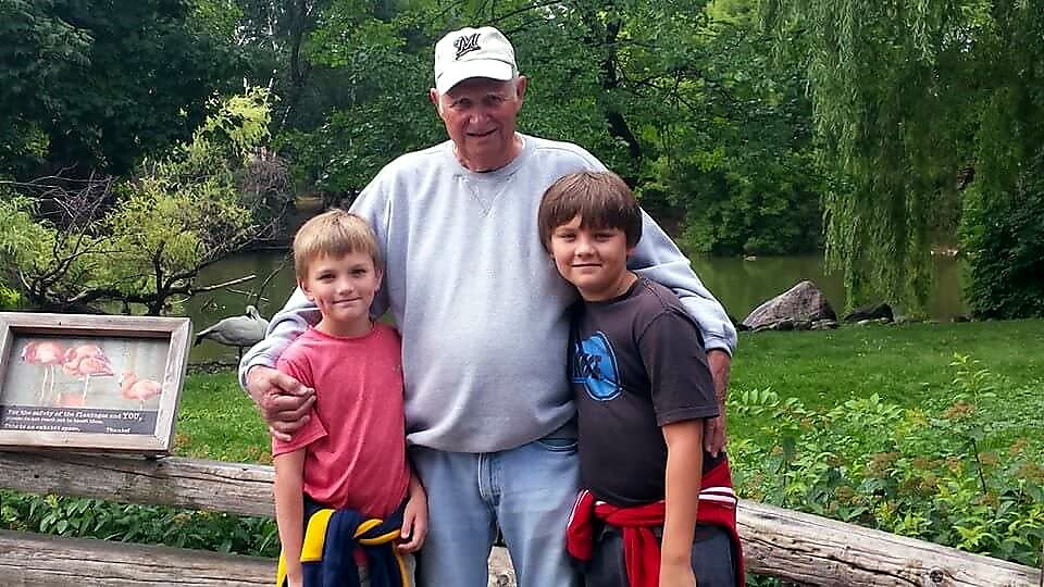 Older man in a cap stands with two young boys in a park, smiles, arms around them.